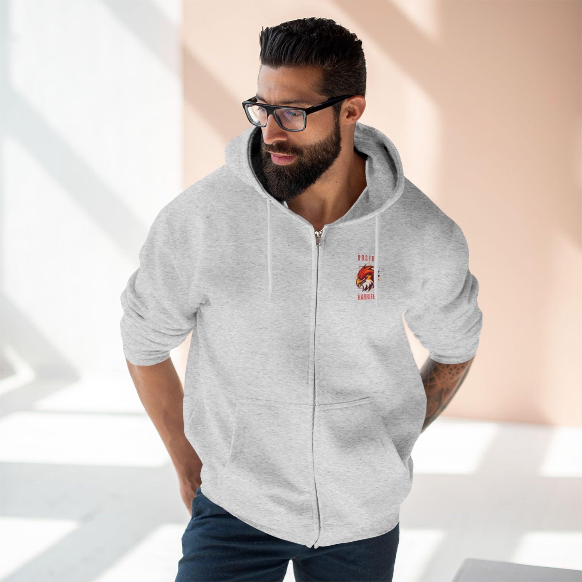 Brown skinned man with dark hair wearing a heather grey hoodie with red Boston Harriers logo on left chest pocket against a peach background