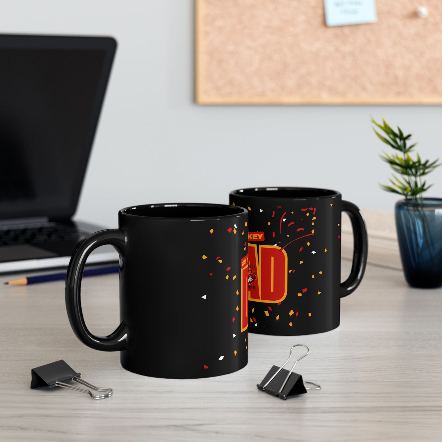 Side view of two black mugs with red and yellow '#1 Hockey Dad' text, Boston Harrier's logo and confetti design on a desk with laptop and office supplies