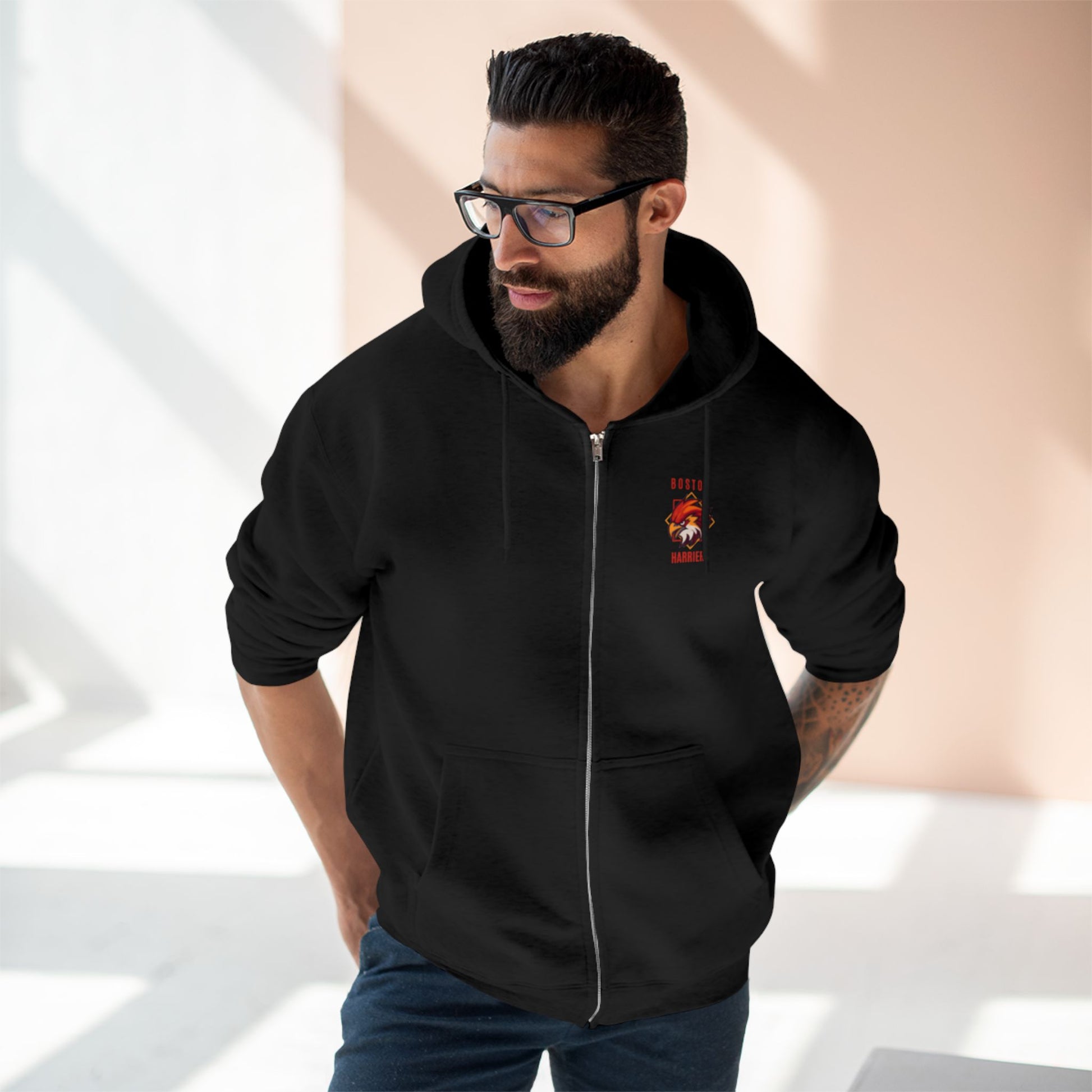 Brown skinned man with dark hair wearing a black hoodie with red Boston Harriers logo on left chest pocket against a peach background
