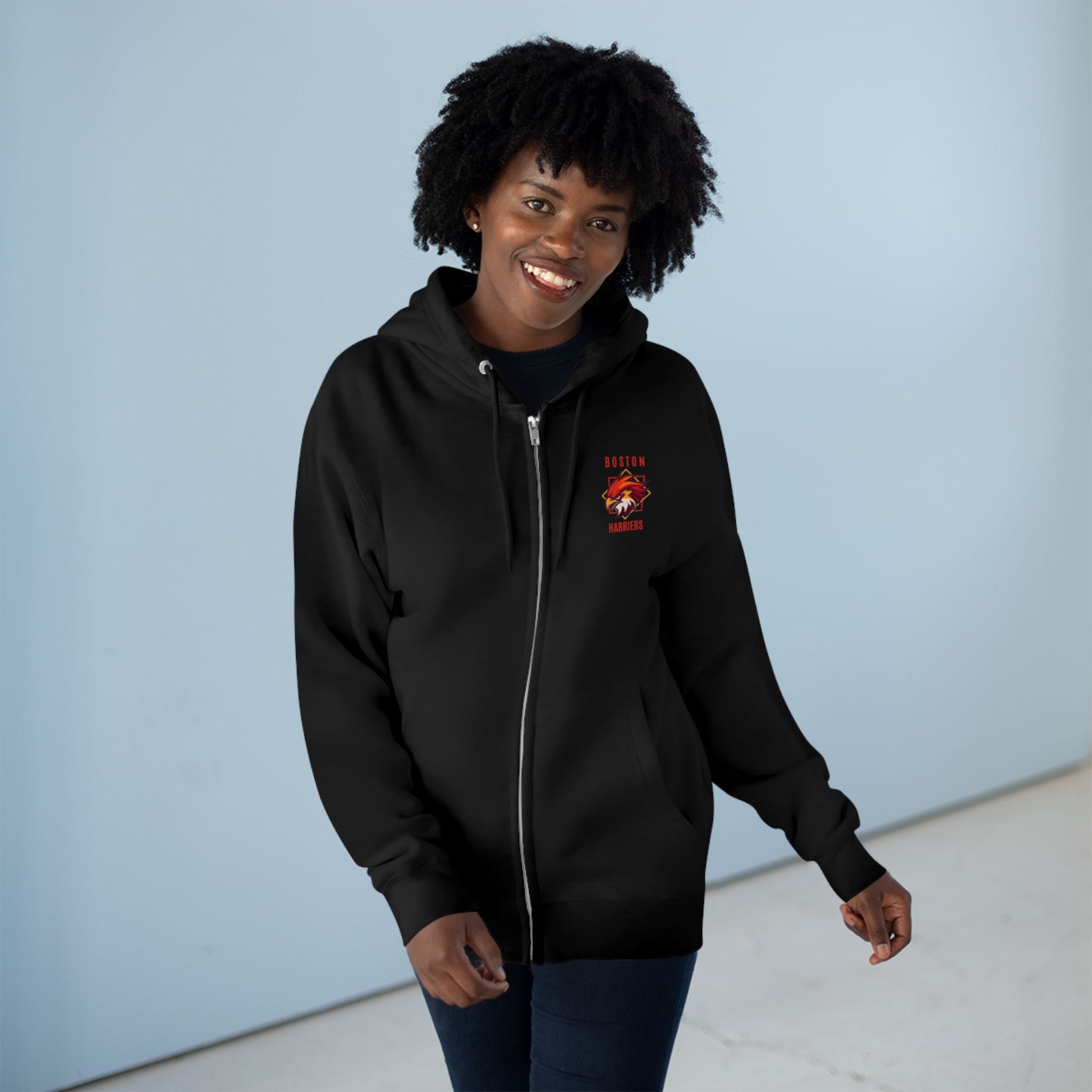 Dark brown skinned woman with natural hair wearing a black hoodie with a white hoodie red Boston Harriers logo against a light blue background