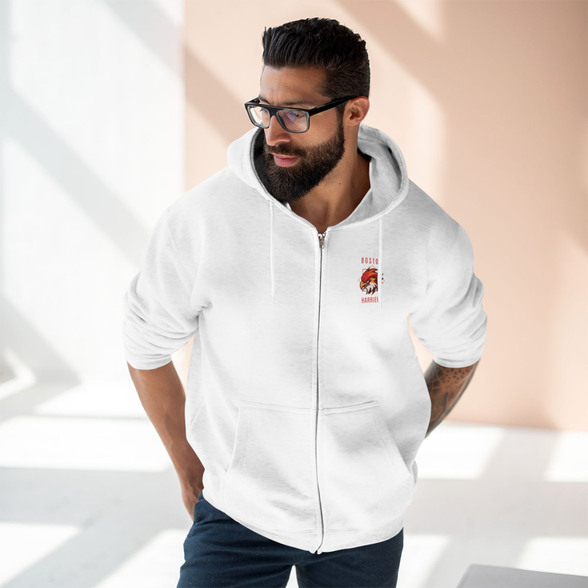 Brown skinned man with dark hair wearing a white hoodie red Boston Harriers logo on left chest pocket against a peach background