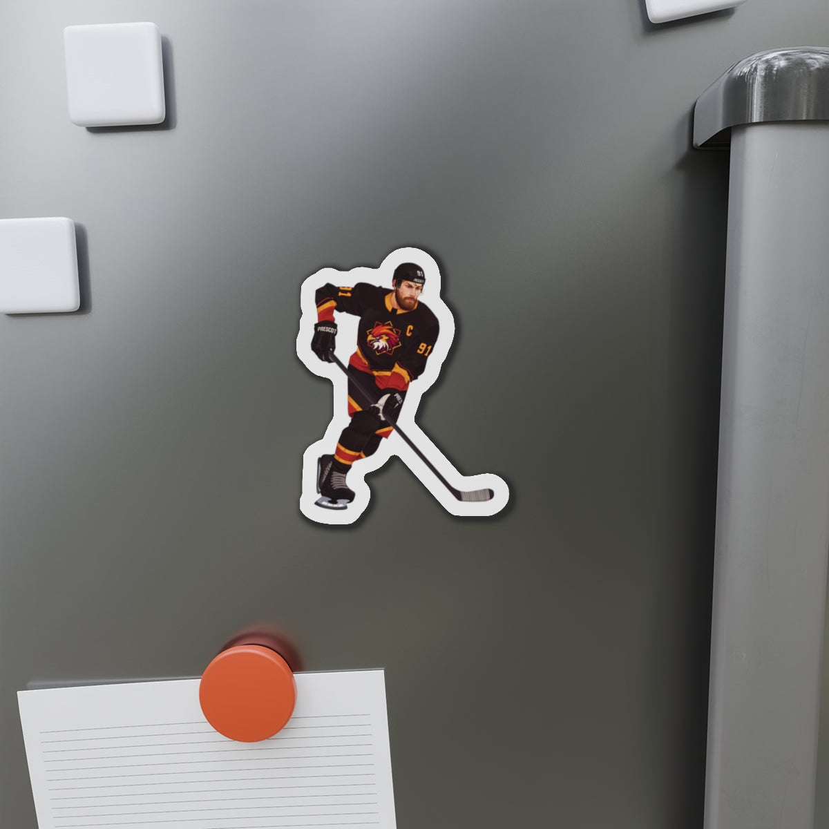 Illustration of a hockey player in a black, red, and gold uniform with a hawk on the chest and C above it. Depicting Connor O'Shea from mm hockey romance Goaltender Interference Book #1 in Brigham Vaughn's Scoring Chances Series. Displayed  refrigerator with other magnets and a paper holder.