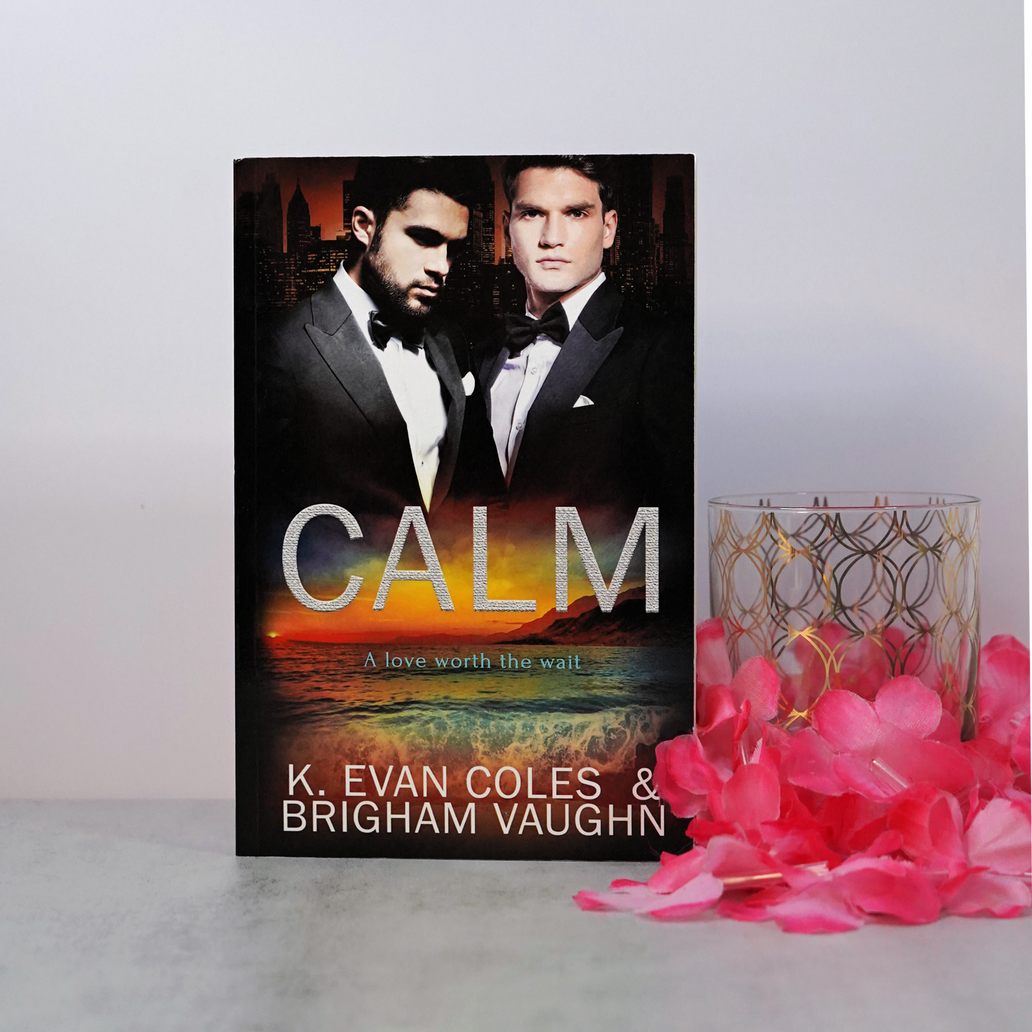 Signed paperback book titled 'Calm' by K. Evan Coles & Brigham Vaughn with two men in tuxedos on the cover, placed on a surface with pink flowers and a highball glass.