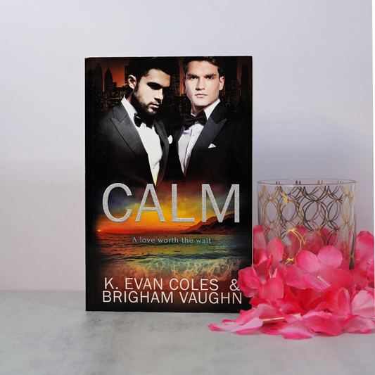 Signed paperback book titled 'Calm' by K. Evan Coles & Brigham Vaughn with two men in tuxedos on the cover, placed on a surface with pink flowers and a highball glass.