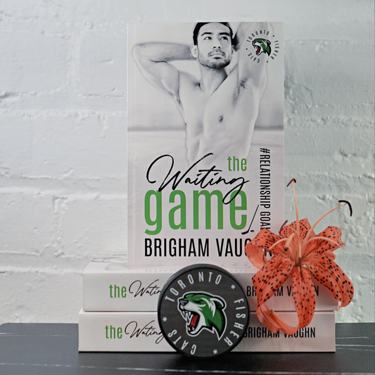 Signed paperback book titled 'The Waiting Game' by Brigham Vaughn with a sports team logo and an orange flower against a white brick wall background.