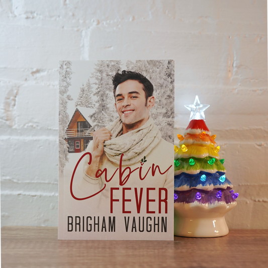 Signed paperback book titled 'Cabin Fever' by Brigham Vaughn with a small, rainbow ceramic Christmas tree on a wooden surface.