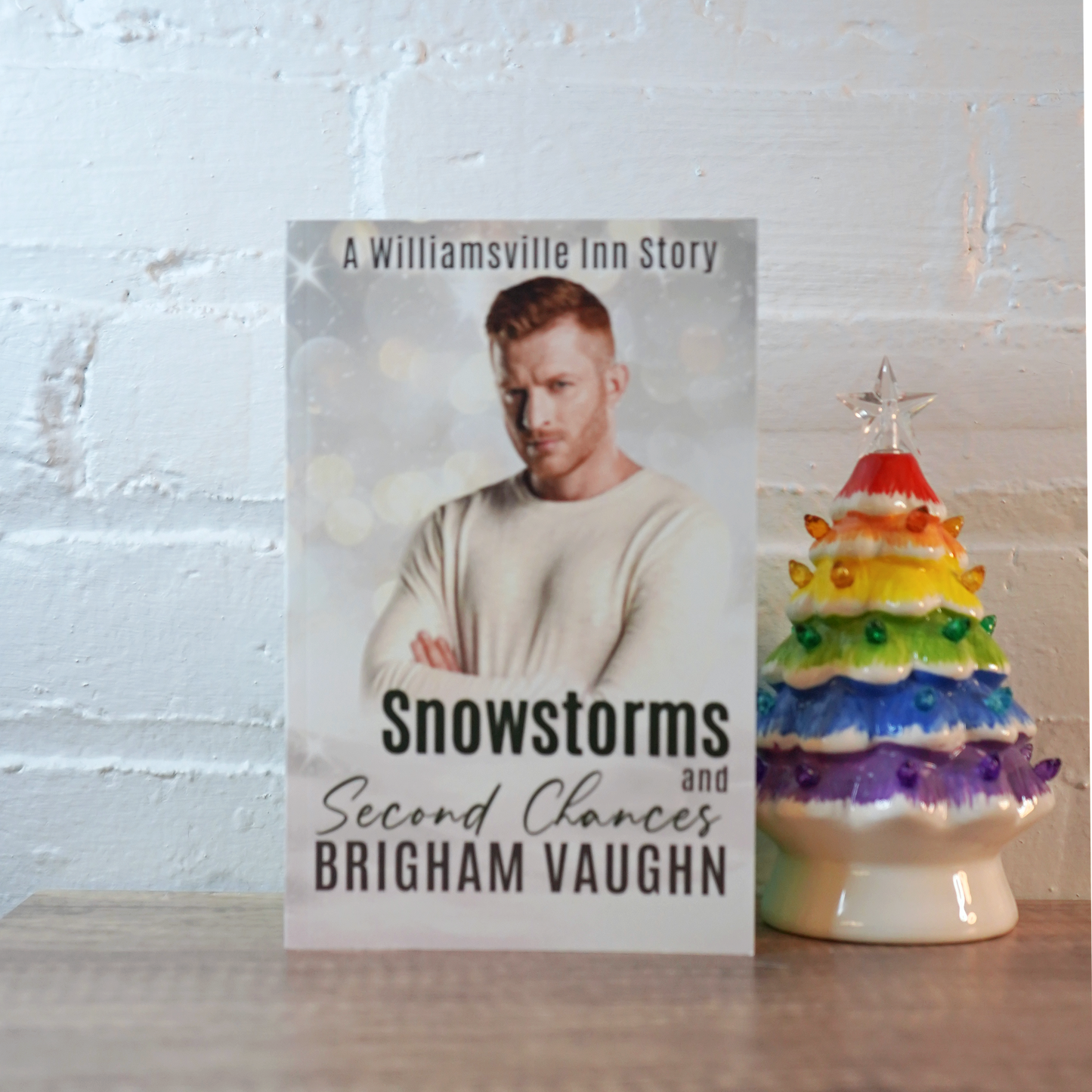 Signed paperback book titled 'Snowstorms and Second Chances' by Brigham Vaughn with a rainbow ceramic Christmas tree on a wooden surface.