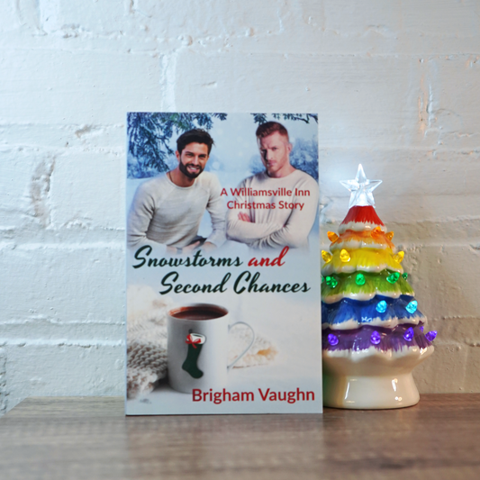 A signed paperback book titled 'Snowstorms and Second Chances' by Brigham Vaughn with a rrainbow ceramic Christmas tree  on a wooden surface.