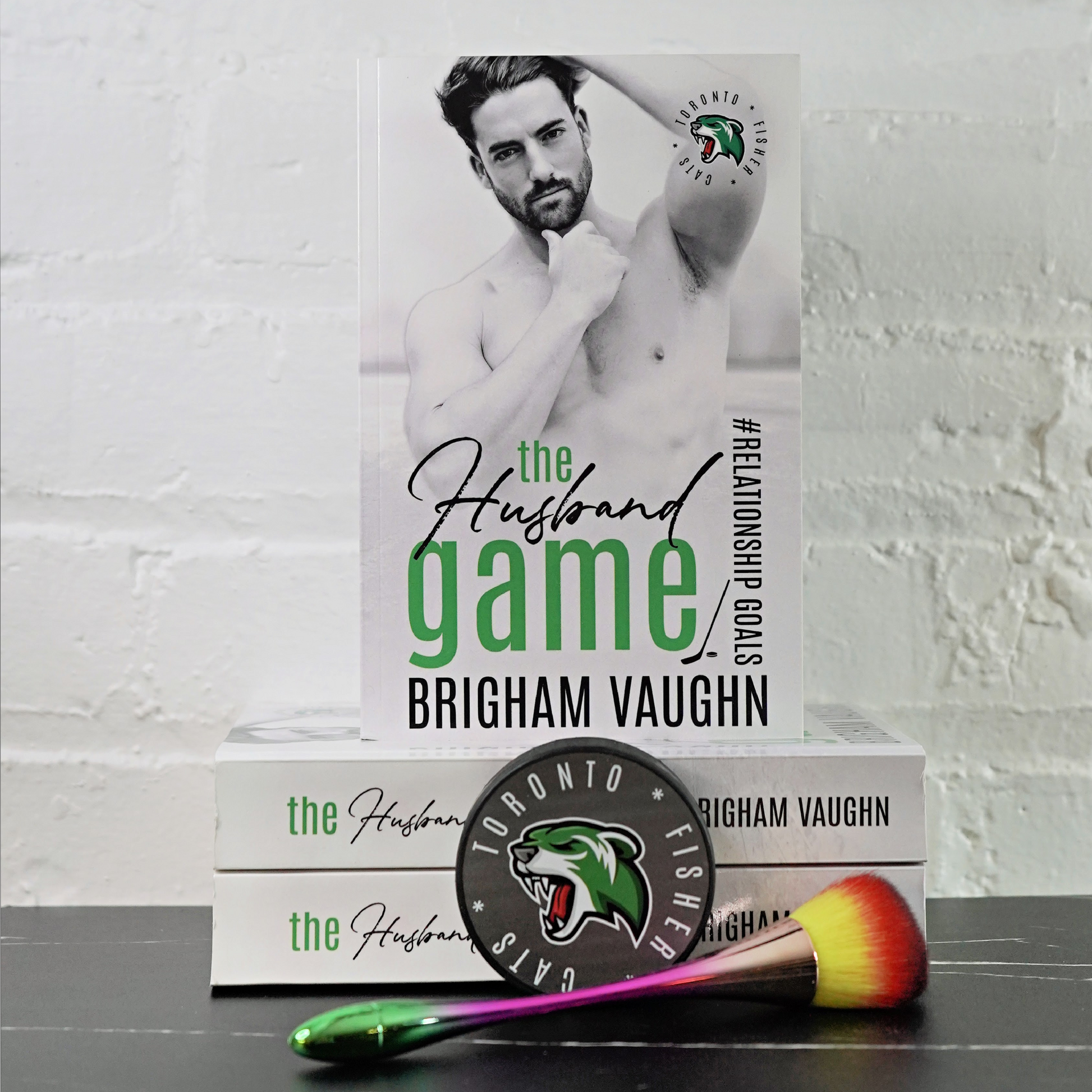 Signed paperback book titled 'The Husband Game' by Brigham Vaughn with a makeup brush and hockey puck on a white brick wall background.
