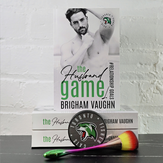 Signed paperback book titled 'The Husband Game' by Brigham Vaughn with a makeup brush and hockey puck on a white brick wall background.