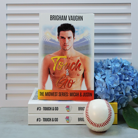 Stack of signed paperback books titled 'Touch & Go' by Brigham Vaughn with a baseball and flowers on a white surface.