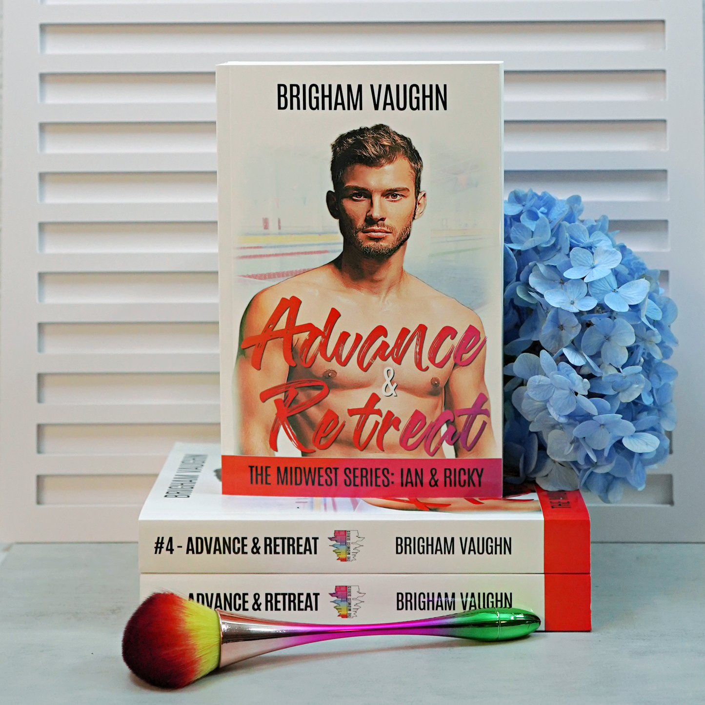 Stack of signed paperback books titled 'Advance & Retreat' by Brigham Vaughn with blue flowers and a makeup brush on a white background.