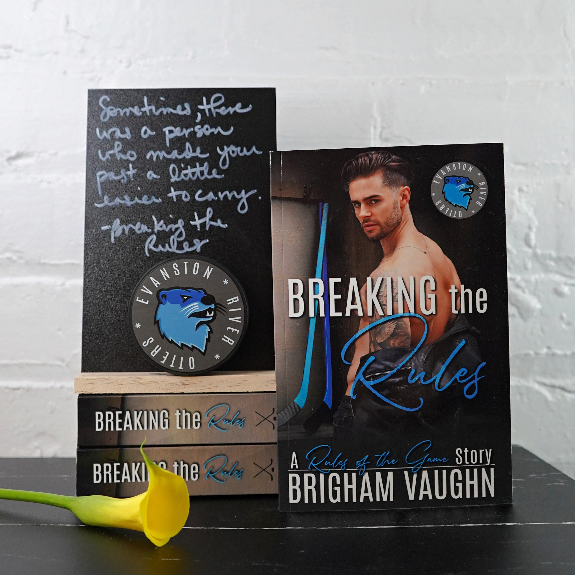 Signed paperback book titled 'Breaking the Rules' by Brigham Vaughn with a quote card and yellow flower against a white brick wall background.