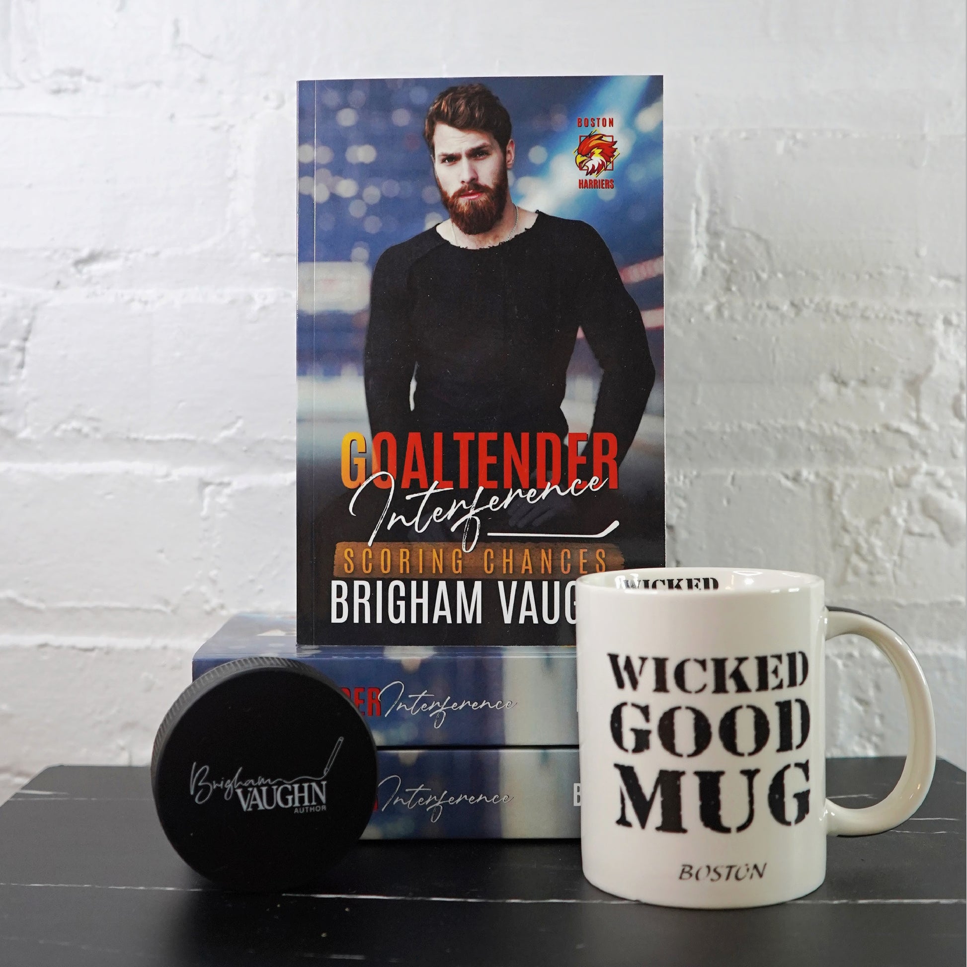Signed paperback book titled 'Goaltender Interference' by Brigham Vaughn with a mug and hockey puck on a white brick wall background.