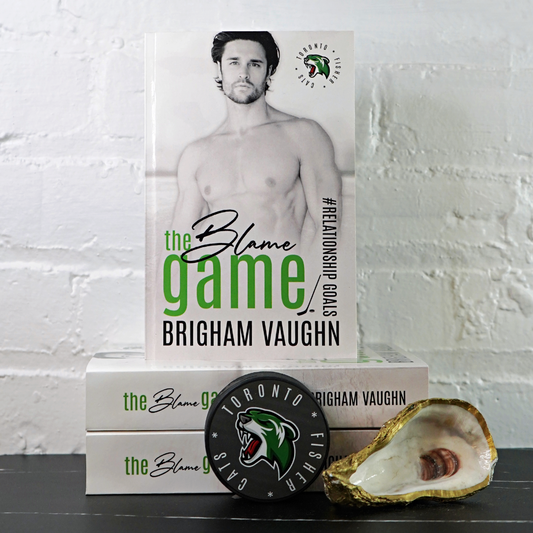 Signed paperback book titled 'The Blame Game' by Brigham Vaughn with a hockey puck and oyster shell against a white brick wall background.