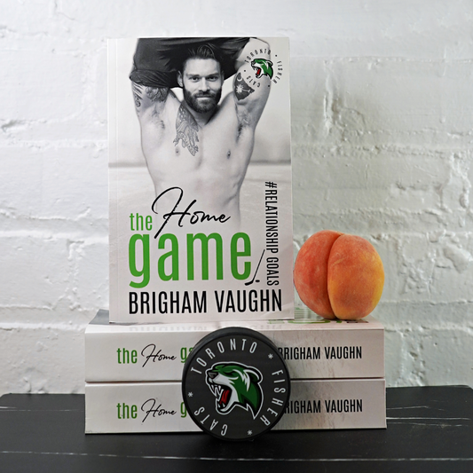 Stack of signed paperback books titled 'The Home Game' by Brigham Vaughn with a hockey puck and a peach against a white brick wall background.