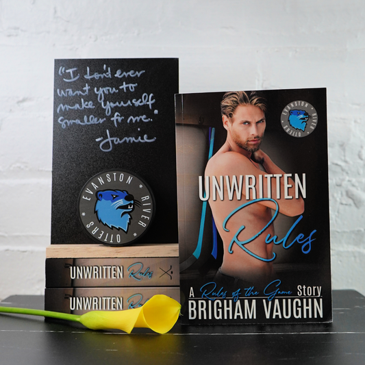 Signed paperback books titled 'Unwritten Rules' by Brigham Vaughn with a stack of books and a yellow flower against a white brick wall background.